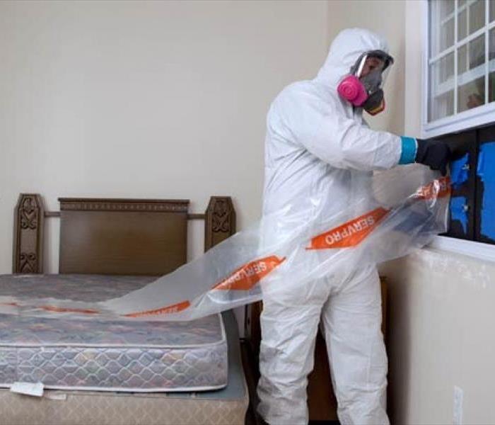 SERVPRO technician in white bio suit with respirator installing negative pressure out of window of room with a bed in it